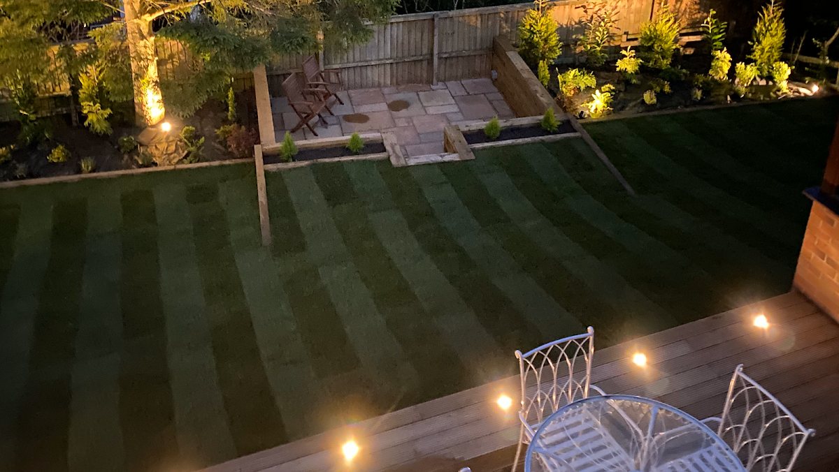 A garden at night lit by fitted garden lighting, with decking, a striped lawn and sleeper terracing
